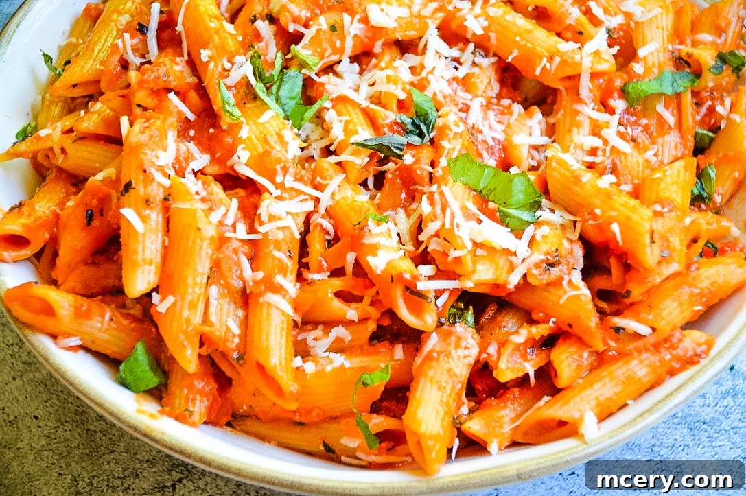 A large bowl of Penne Alla Vodka is shown from above, with the pasta glistening in its creamy tomato sauce. It's generously topped with freshly grated Parmesan cheese and vibrant green basil leaves, positioned invitingly on a wooden surface.