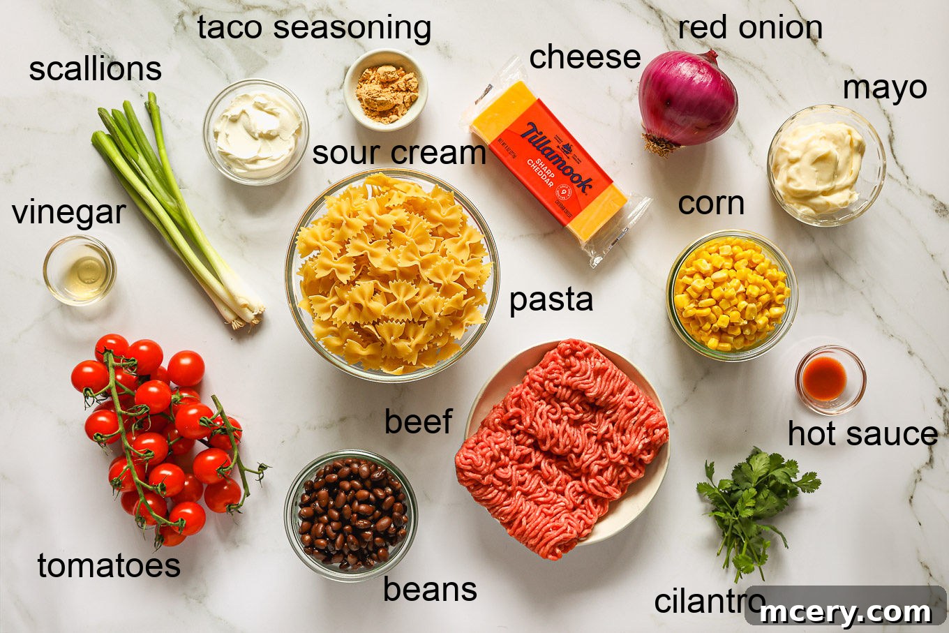 A flat lay photograph showcasing all the fresh and pantry ingredients laid out for making Cowboy Pasta Salad, including farfalle pasta, ground beef, corn, black beans, cherry tomatoes, red onion, cheddar cheese, cilantro, mayonnaise, sour cream, and spices.