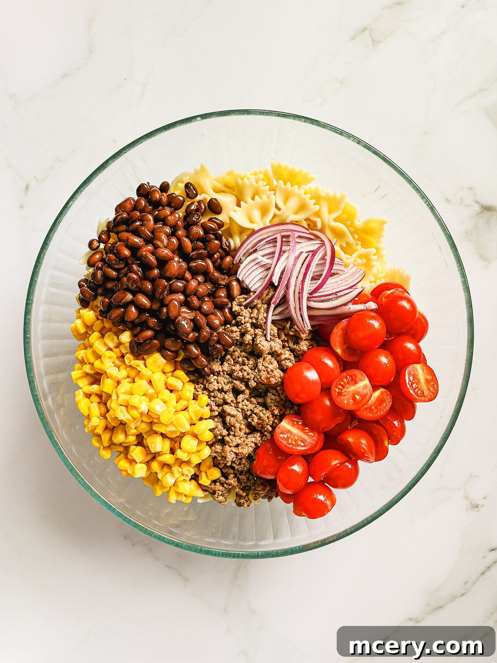 A large glass bowl filled with cooked pasta, seasoned ground beef, black beans, corn, and cherry tomatoes, ready for the dressing and final ingredients of the Cowboy Pasta Salad.