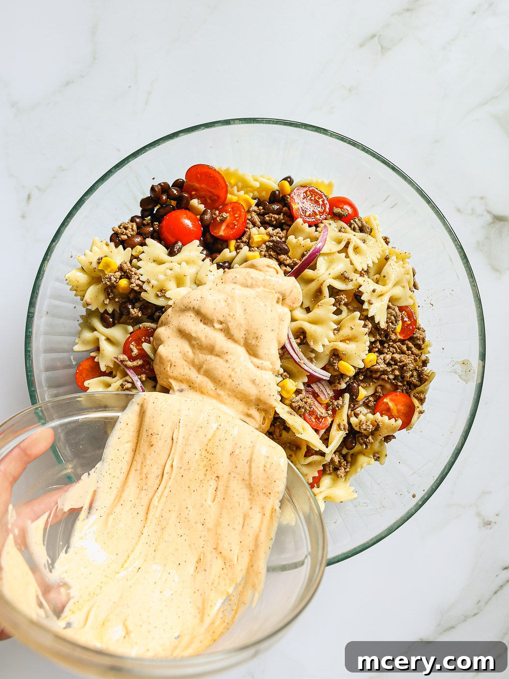 The creamy dressing being poured over the mixed pasta salad ingredients in a glass bowl, ready for tossing to combine all flavors.