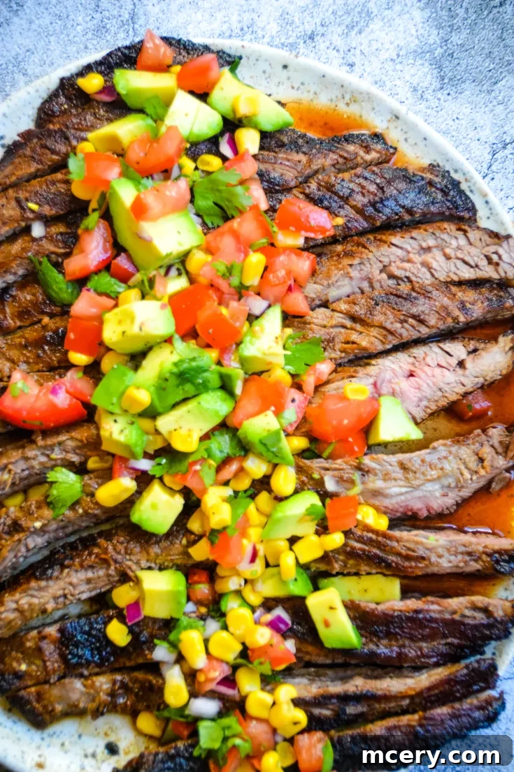 A plate of grilled Spice Rubbed Flank Steak & Avocado Salsa, consisting of a colorful mixture of diced tomatoes, avocado, corn, red onions, and cilantro, placed on a light gray surface. The steak is sliced thinly and the vibrant toppings contrast with the rich, dark grill marks on the meat.
