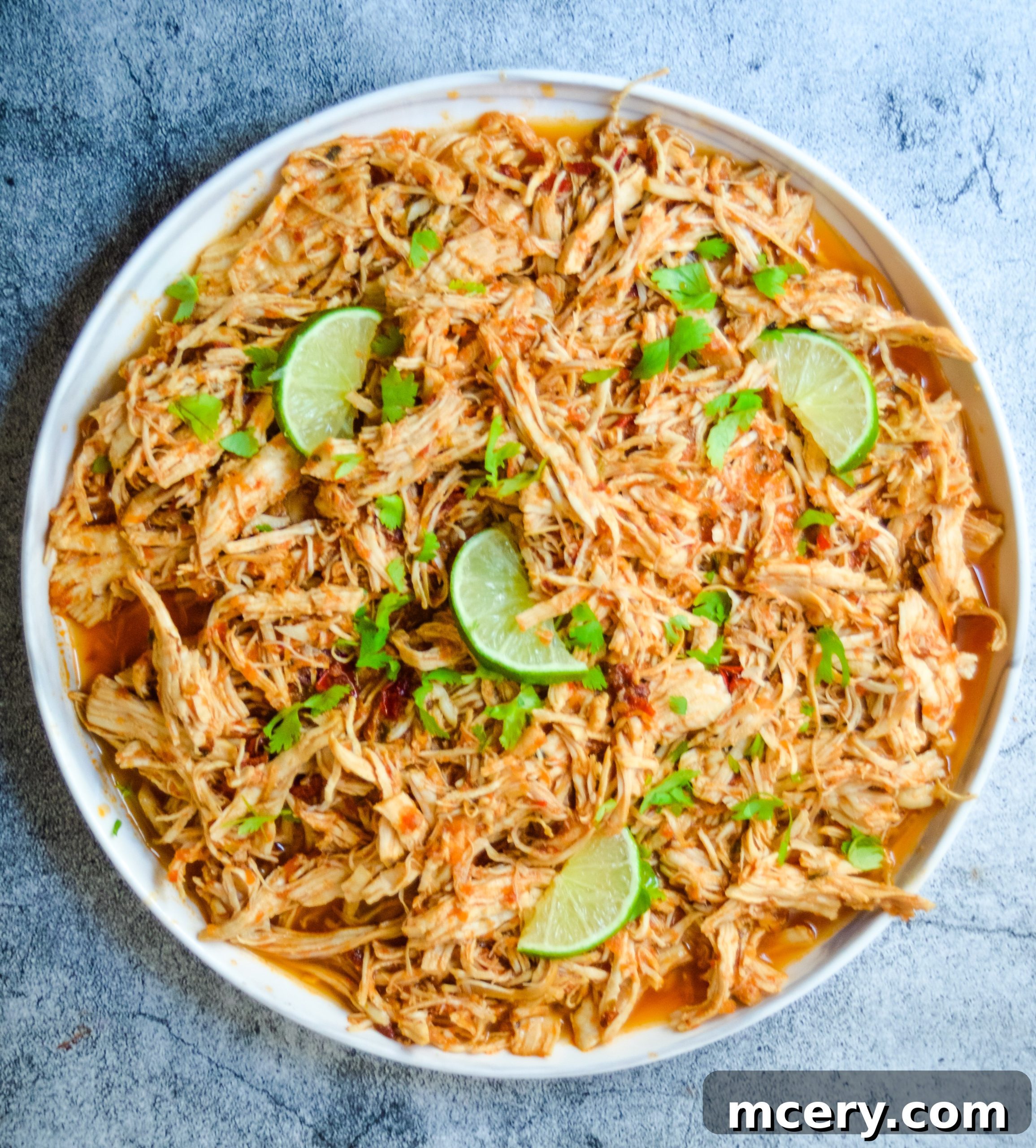 Speedy Instant Pot Mexican Pulled Chicken 4 A close-up of a serving of Instant Pot Shredded Mexican Chicken, garnished with fresh cilantro and lime wedges, presented on a white plate. The chicken is visibly tender and coated in a rich, flavorful sauce, ready to be served.