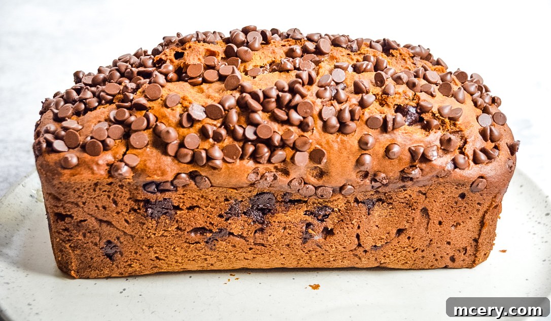 A large loaf of freshly baked Pumpkin Chocolate Chip Bread, partially sliced, displayed on a wooden cutting board with a knife beside it.
