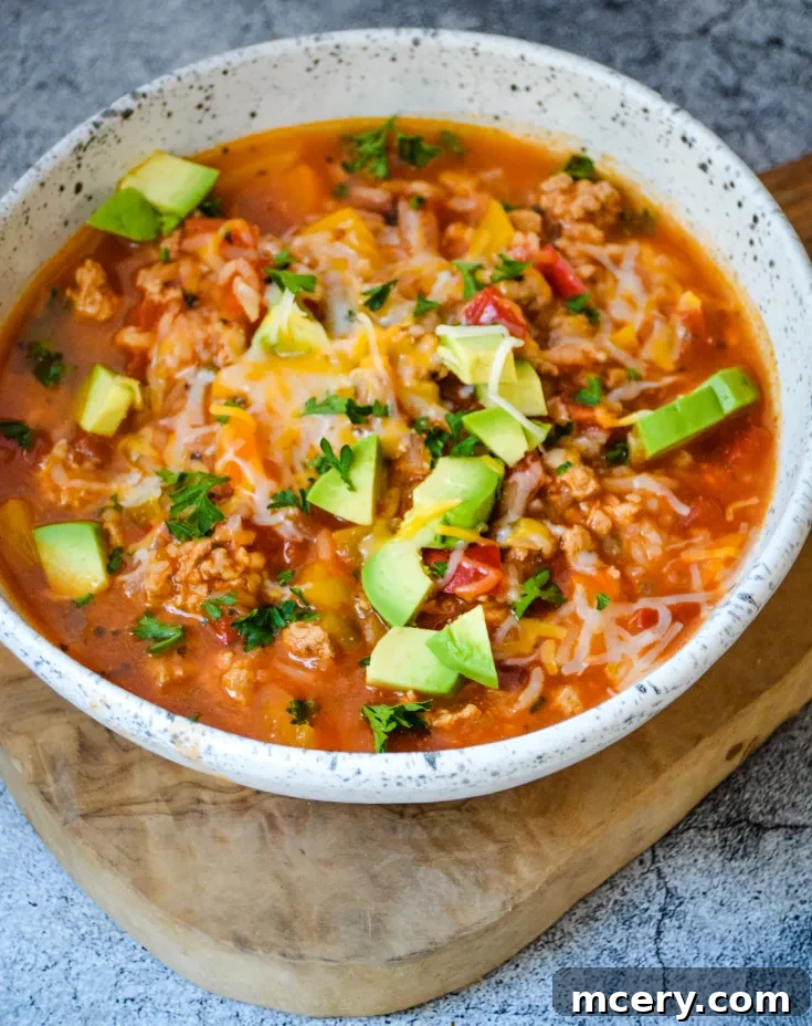Savory Stuffed Bell Pepper Pot 7 A savory bowl of Stuffed Pepper Soup featuring a blend of ground meat, tomatoes, diced avocados, shredded cheese, and chopped herbs, served in a white speckled bowl placed on a wooden cutting board. The dish is colorful and garnished with fresh parsley.
