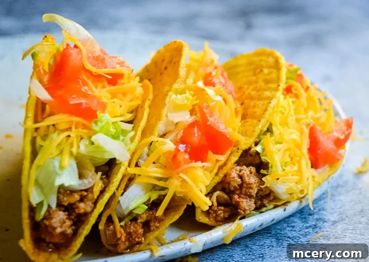 A white rectangular dish filled with crispy Instant Pot Ground Turkey Tacos. Each taco is packed with a mix of seasoned ground turkey meat, shredded cheese, diced tomatoes, crisp lettuce, and topped with fresh avocado slices. The dish is set against a light gray background, highlighting the vibrant colors and inviting texture of the tacos.