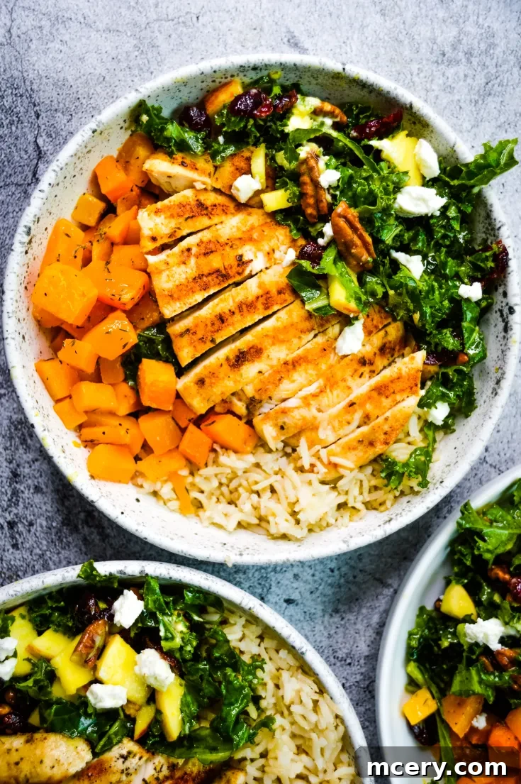 Fall Harvest Bowls, featuring grilled chicken slices on a bed of rice, accompanied by roasted sweet potatoes, kale salad, pecans, cranberries, and crumbled feta cheese. The bowl sits on a textured surface, with a portion of two similar bowls visible in the corners.