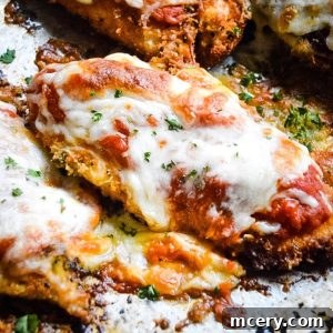 A close-up of Crispy Chicken Parmesan on a metal baking sheet. The breaded chicken cutlets are topped with melted mozzarella cheese and marinara sauce. Chopped parsley is sprinkled on top for garnish. The edges are crispy and slightly browned.