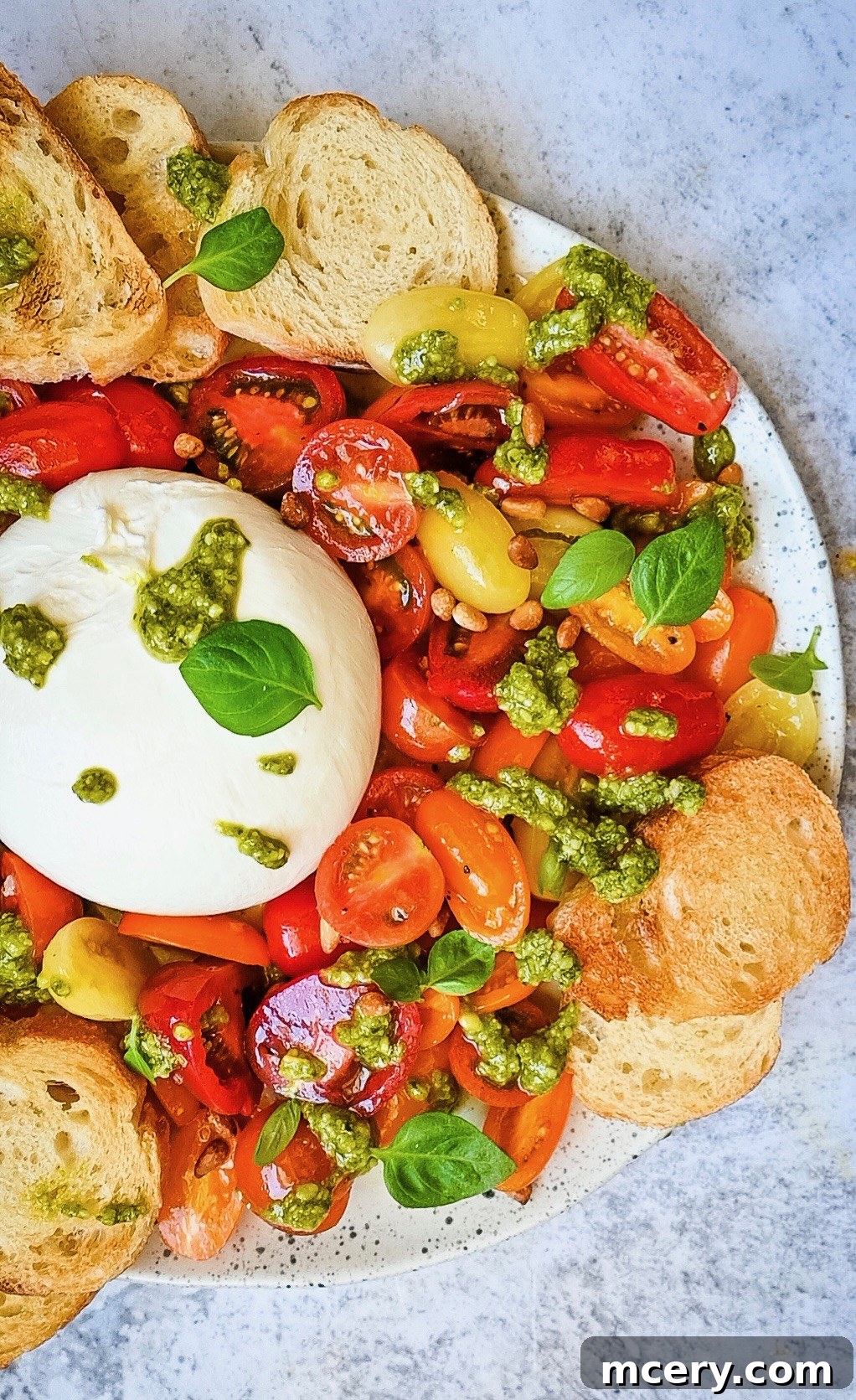 Velvety Burrata with Ripe Tomatoes and Fragrant Pesto 2 A beautifully plated Fresh Tomato and Burrata Salad with Pesto, showcasing a vibrant array of red and yellow cherry tomatoes, a generous ball of creamy burrata, and rich green pesto, artfully arranged on a white plate. Toasted garlic bread slices are visible around the edges, inviting a delightful culinary experience.