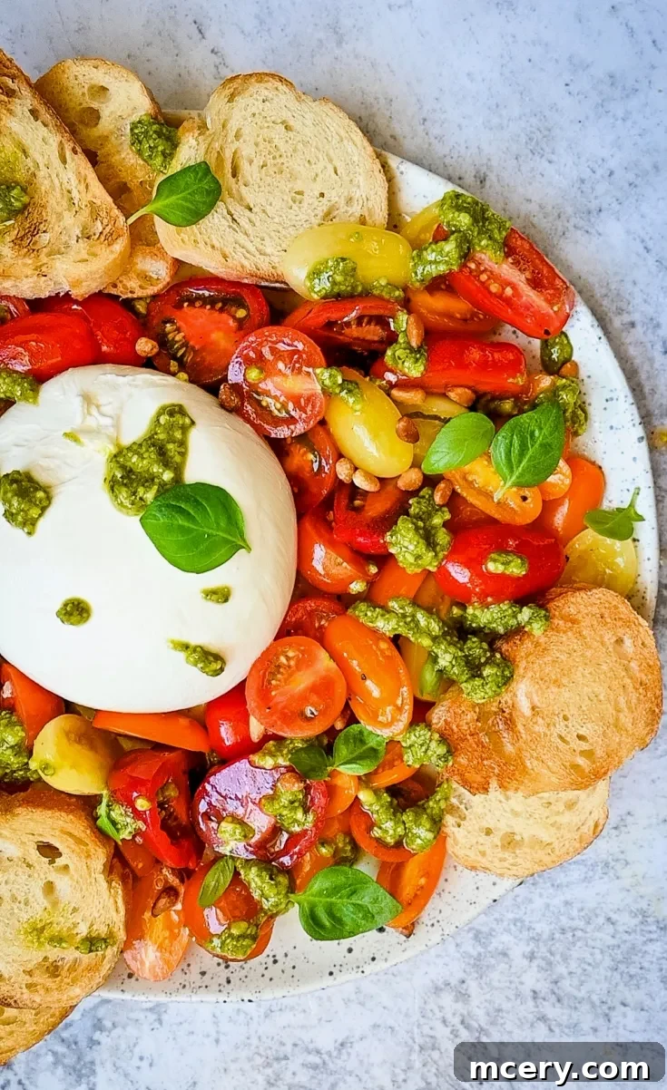 A colorful plate featuring Fresh Tomato and Burrata Salad with Pesto showing a variety of cherry tomatoes, yellow tomatoes, and red tomatoes, topped with a ball of fresh burrata, dollops of green pesto, fresh basil leaves, and surrounded by slices of toasted bread.