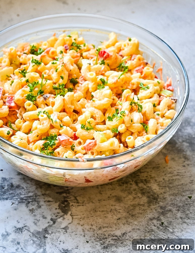 A bowl of creamy macaroni salad, garnished with fresh dill and finely chopped vegetables, ready to be served. The dressing is perfectly coating each noodle and vegetable piece.