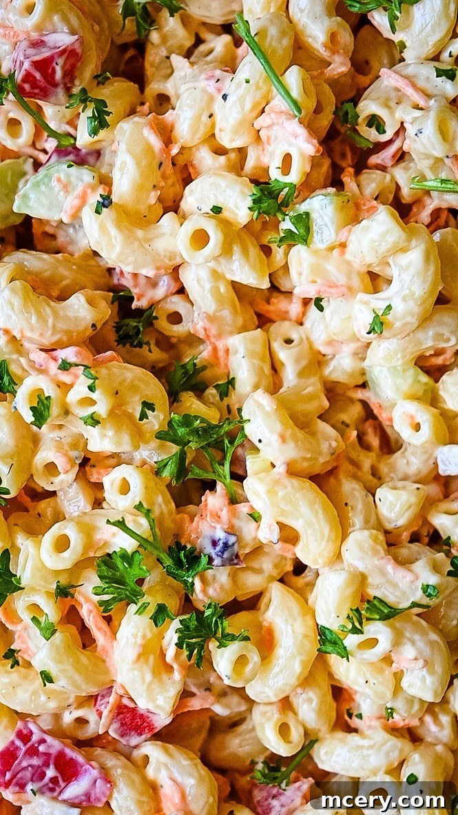 Close-up of a creamy macaroni salad mixed with chopped vegetables like red bell peppers, cucumbers, and carrots, garnished with fresh parsley. The pasta is coated evenly with a thick, white dressing, giving it a vibrant and appetizing look.