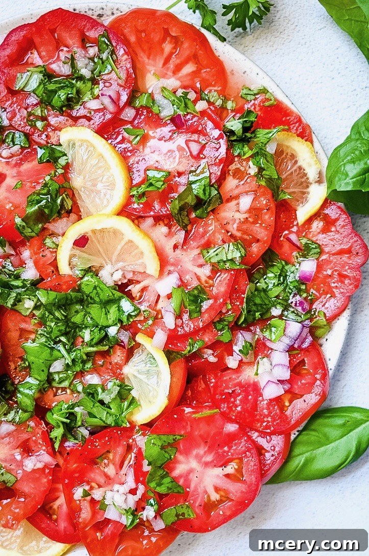 A close-up of a fresh salad featuring vibrant Marinated Tomato slices topped with finely chopped green herbs, red onions, and thin lemon slices, served on a white plate. Fresh parsley and basil leaves are visible in the background.