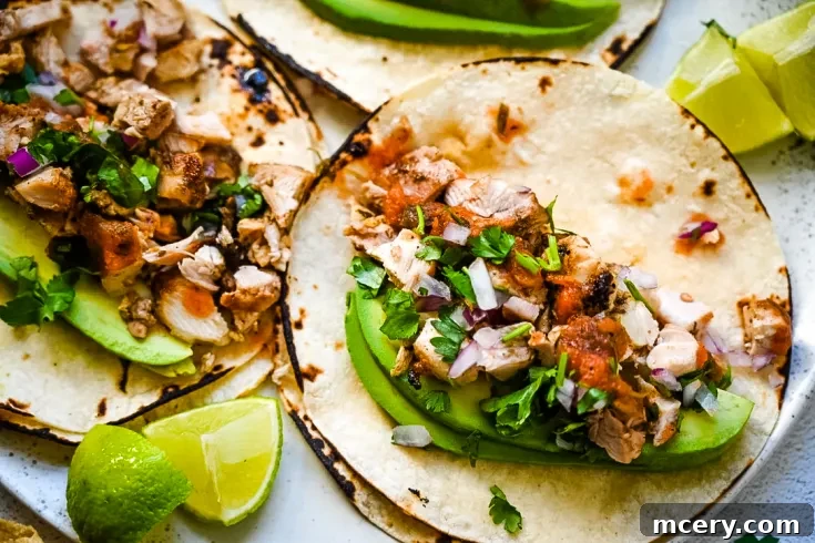 A close-up of Easy Grilled Chicken Street Tacos with corn tortillas filled with slices of avocado, shredded cooked meat, diced red onions, chopped fresh cilantro, and a drizzle of red salsa. The taco is on a white plate with lime wedges visible in the background.