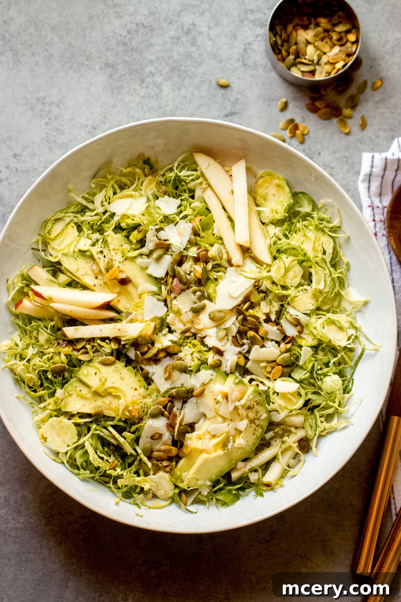 Harvest Apple Brussels Sprout Salad 2 shredded Brussels sprouts salad in a white bowl with wooden utensils on the side.