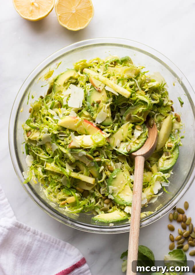 Harvest Apple Brussels Sprout Salad 7 tossed shaved Brussels sprouts with apples, avocado, and dressing in a glass bowl with a wooden spoon.