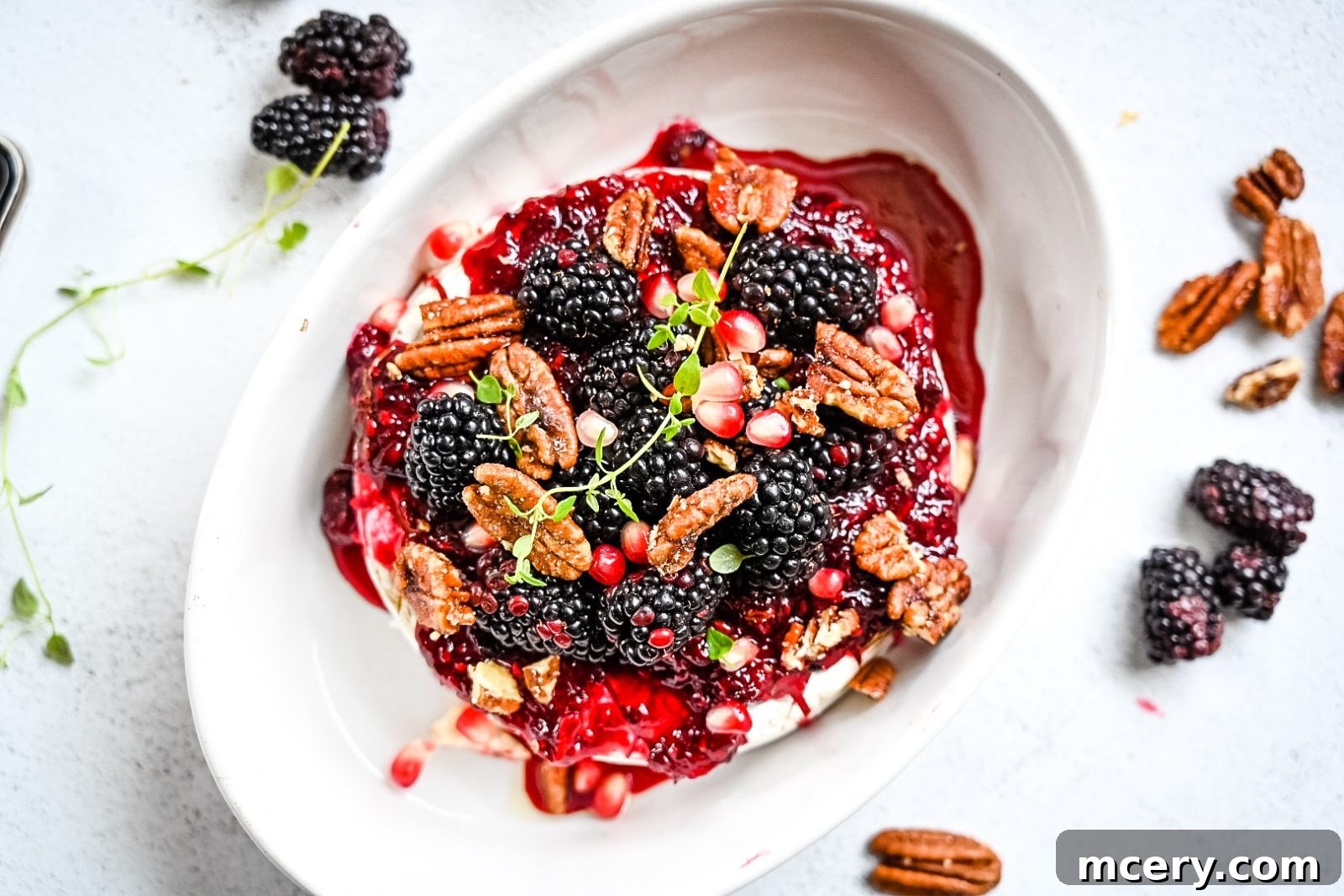 A beautifully presented baked brie, glistening with homemade blackberry compote and adorned with fresh blackberries, candy pecans, and thyme leaves. The cheese sits in a rustic baking dish, ready to be served.