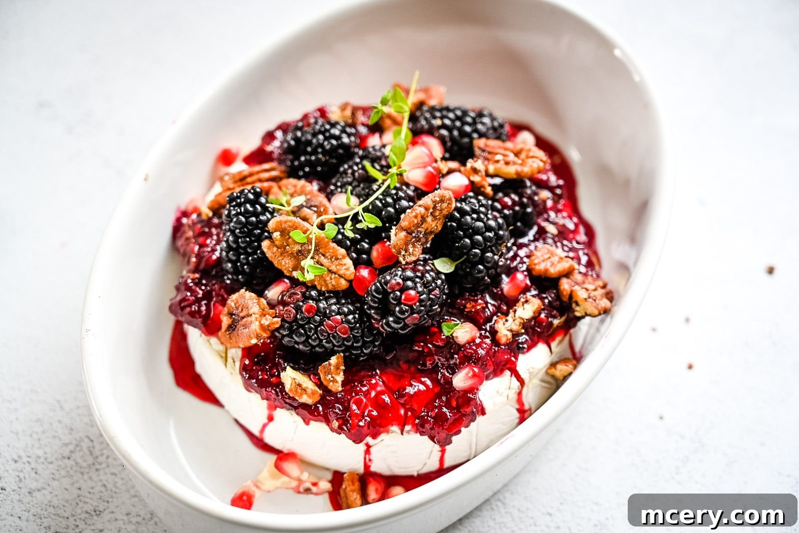 The finished Blackberry Baked Brie, adorned with fresh blackberries, pomegranate seeds, candy pecans, and thyme, in a rustic ceramic baking dish. The rich colors are inviting and appetizing.
