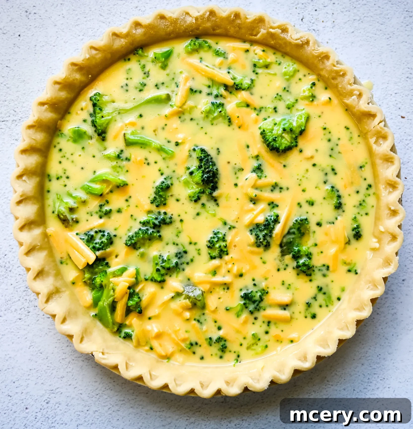 Ingredients for a broccoli cheddar quiche laid out, including a pie crust, fresh broccoli, eggs, and shredded cheddar cheese.