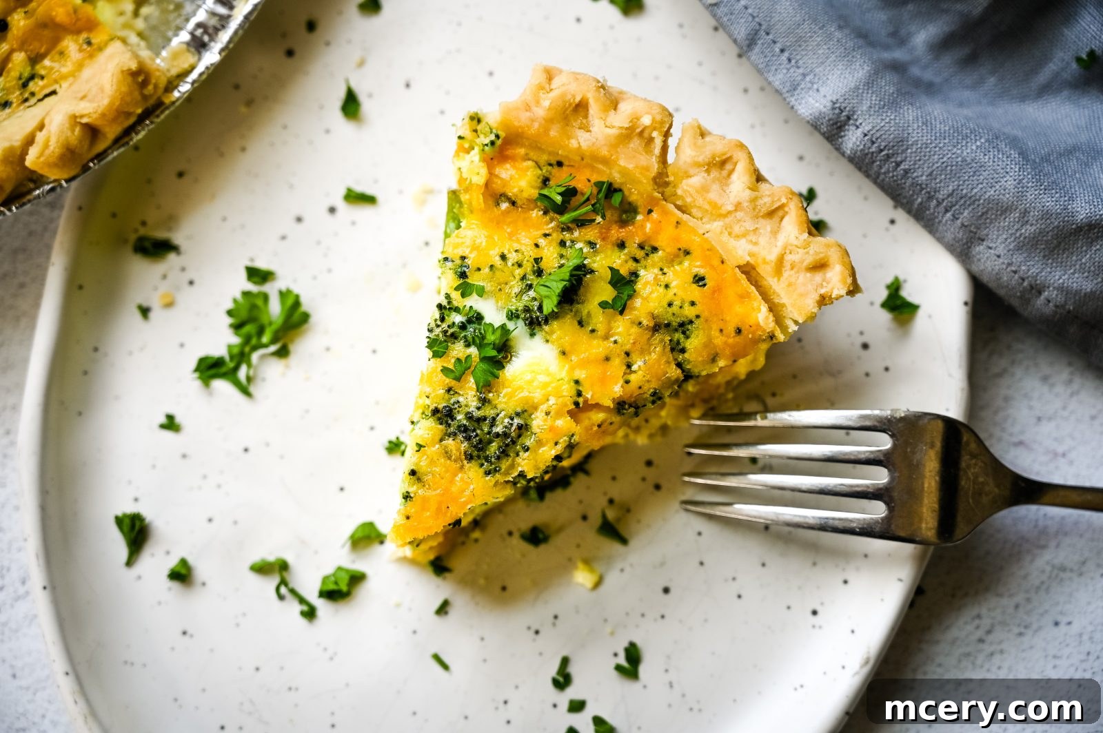 A slice of Easy Broccoli Cheddar Quiche on a white plate with a fork beside it. The quiche has a golden crust, visible broccoli florets, and a creamy, cheesy filling. Fresh parsley is sprinkled on top, and a blurred background shows more quiche in a pie tin.