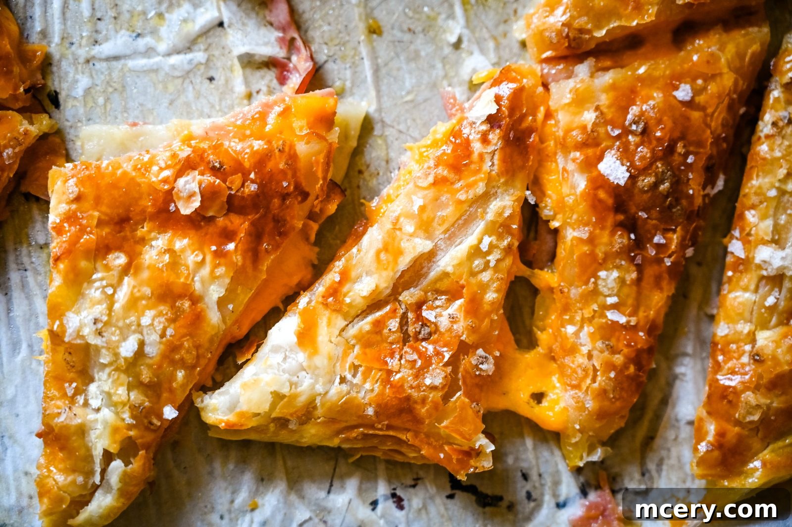 A close-up of freshly baked, golden-brown Ham Cheddar & Apple Puffs. Cut into triangles and sprinkled with coarse salt, arranged neatly on a piece of parchment paper. The pastry looks crispy and flaky, with a shiny, slightly caramelized surface.