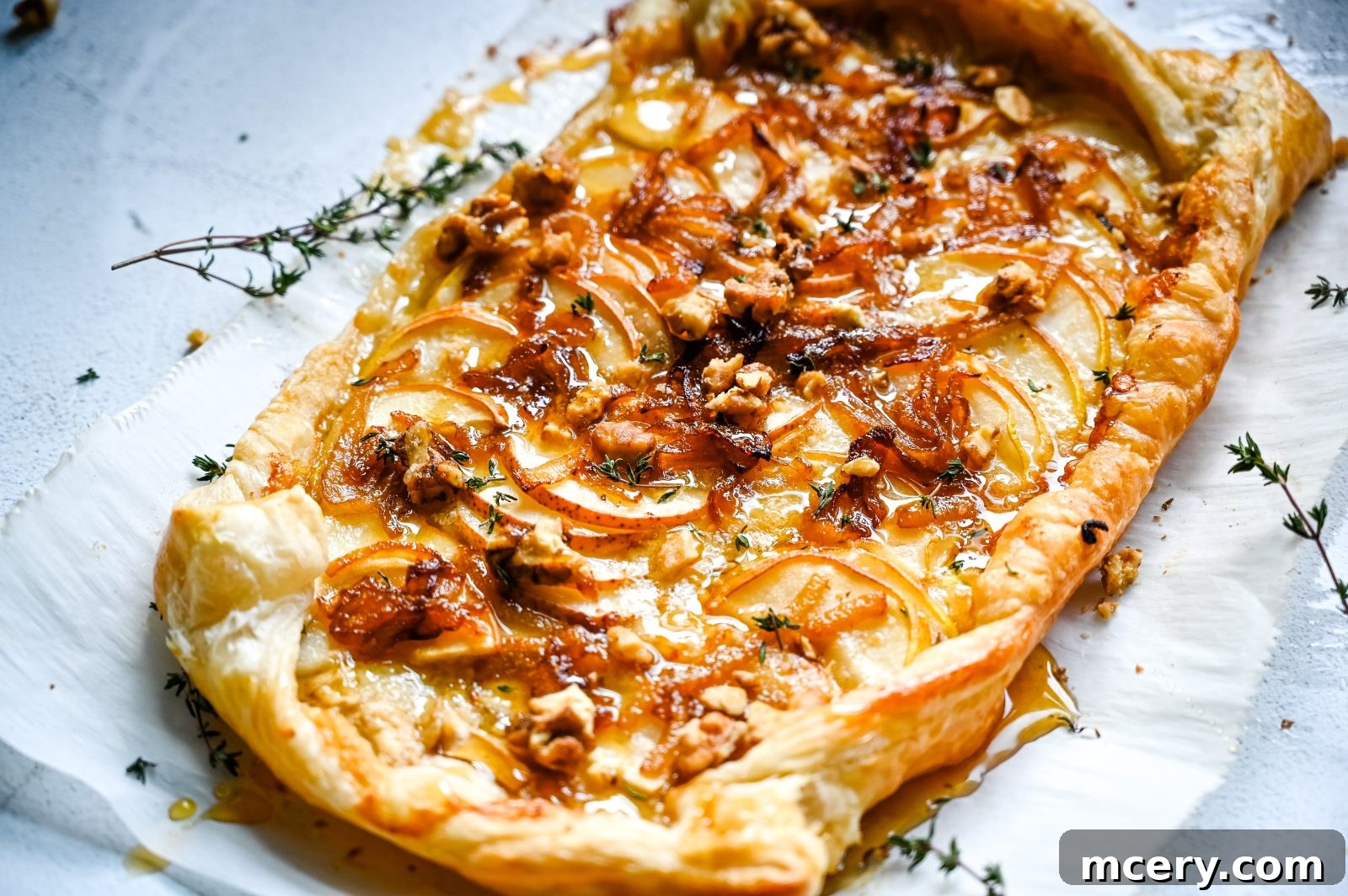 A rectangular Brie and Pear Puff Pastry Tart is topped with caramelized onions, thin slices of pear, chopped walnuts, fresh thyme, and melted cheese. The edges of the pastry are golden and flaky, and the tart is placed on a piece of white parchment paper.