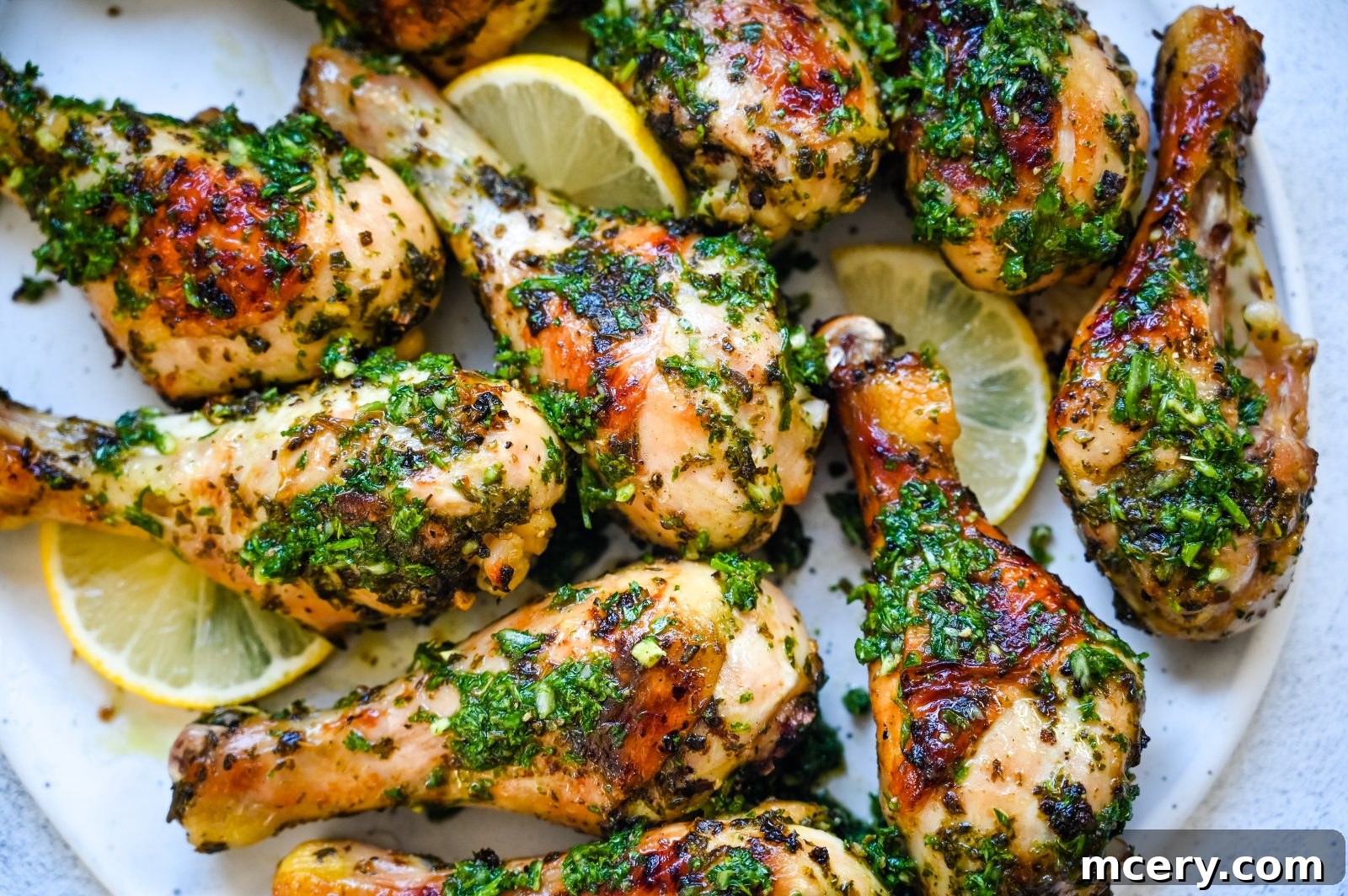 A plate of Delicious Baked Chicken Legs garnished with fresh herbs and lemon slices. The chicken is seasoned with a green herb mixture, and lemon wedges are placed around the edges, all presented on a white plate over a light background.