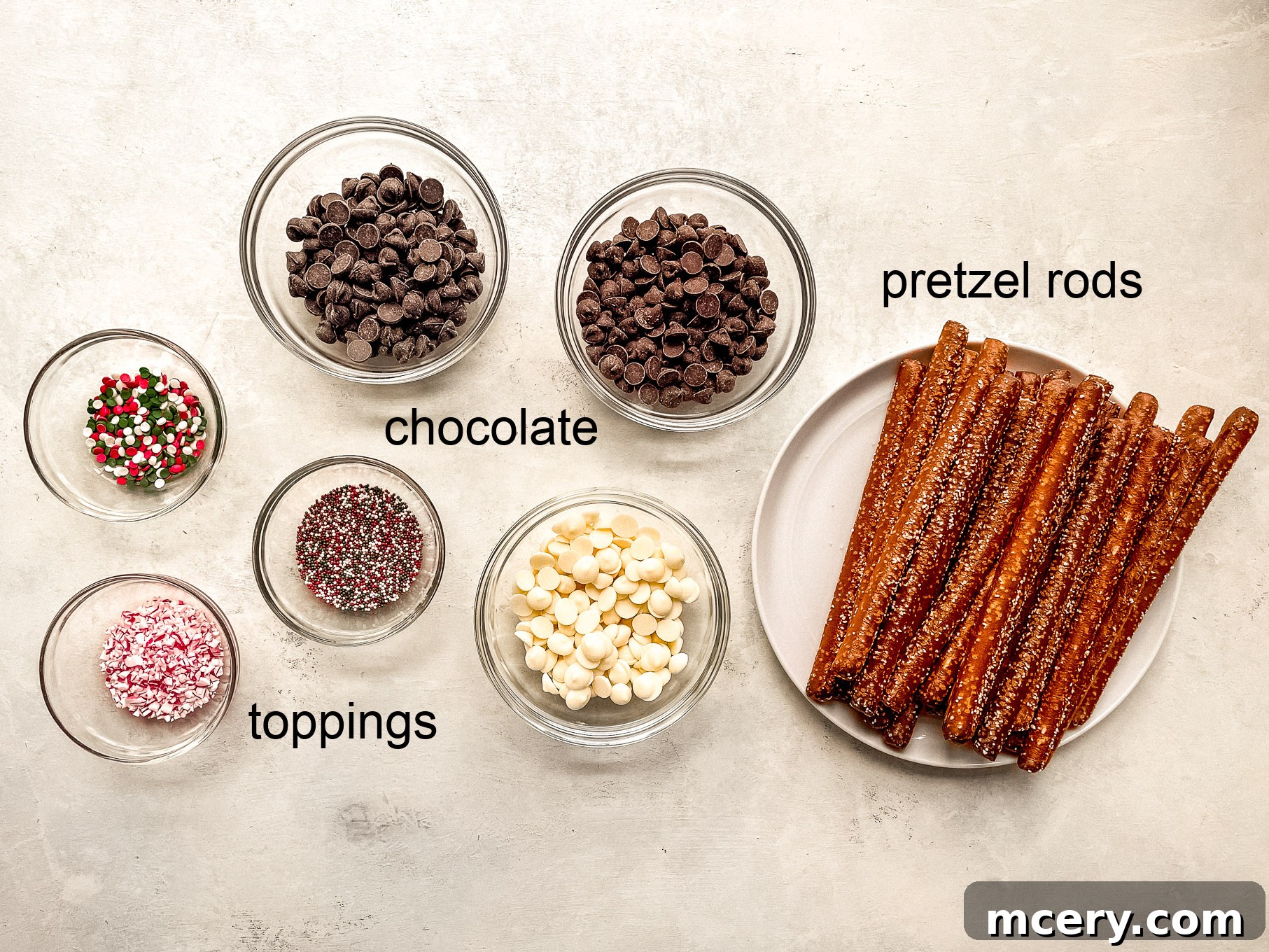Assorted ingredients for making Christmas chocolate pretzels, including pretzel rods, chocolate, and various toppings.