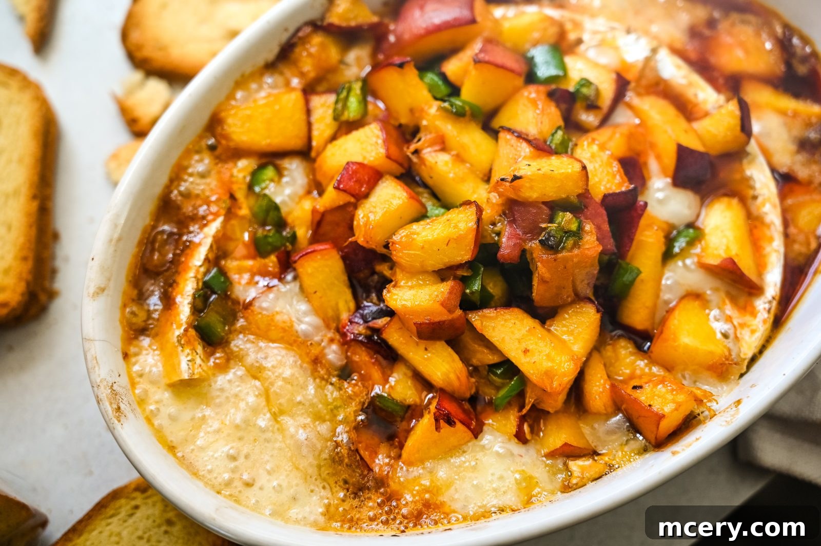 Honey Peach & Chipotle Baked Brie with melted brie topped with diced peaches and jalapeños, presented in a white oval dish against a light background. The cheese is golden and bubbly, creating a vibrant and appetizing appearance.