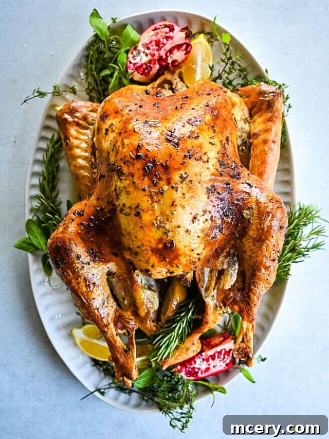 Skip the Brine! Your Easiest, Juiciest Thanksgiving Turkey Ever 2 A perfectly roasted turkey with golden, crispy skin is presented on a white platter, garnished with fresh herbs, lemon wedges, and pomegranate seeds, creating an appetizing display ideal for a festive or holiday meal.
