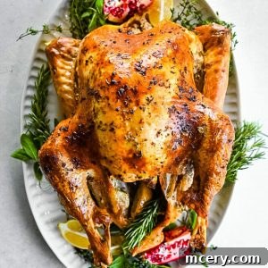 Skip the Brine! Your Easiest, Juiciest Thanksgiving Turkey Ever 13 A perfectly roasted turkey on a white platter is garnished with fresh herbs, lemon wedges, and pomegranate seeds. The turkey has a golden, crispy skin, and the platter is arranged on a smooth, light-colored surface.