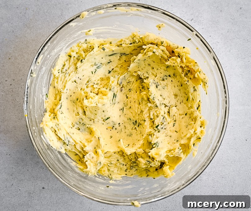 Skip the Brine! Your Easiest, Juiciest Thanksgiving Turkey Ever 6 A clear glass bowl containing a mixture of softened butter blended with finely chopped herbs, placed on a light gray surface. The butter is smooth and creamy, with visible green herb flecks scattered throughout.