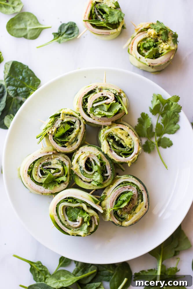Refreshing Cucumber Bites 2 Freshly prepared cucumber roll ups with avocado hummus, ham, and cheese on a white plate