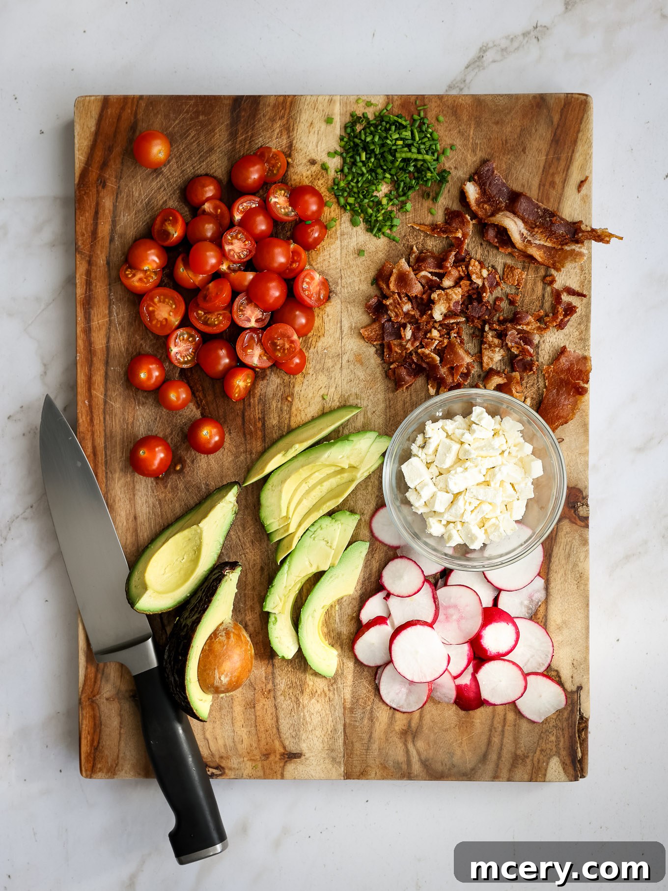 The Ultimate Chopped Wedge Salad 6 A wooden board laden with perfectly sliced and diced ingredients for a chopped wedge salad, featuring halved tomatoes, ripe avocado, crisp radishes, savory bacon, fresh chives, and crumbled feta cheese.