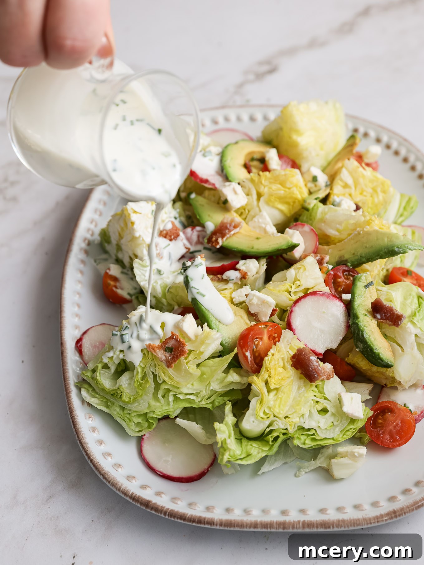 The Ultimate Chopped Wedge Salad 7 A vibrant iceberg lettuce salad on a white platter, being generously drizzled with creamy homemade buttermilk dressing, showcasing the final assembly of this delightful dish.